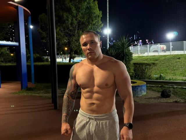 OstenFrey, shirtless, muscular with visible abs, tattoo on right arm, wearing gray pants, black watch, standing in a dark, lit-up park at night, looking forward, slightly sweaty.