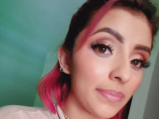 LizBonnet, avec ses cheveux teints en rose, ses sourcils épais, ses longs cils, son rouge à lèvres rose et son sourire discret. Elle porte une petite boucle d'oreille et un collier. Sa peau est claire..