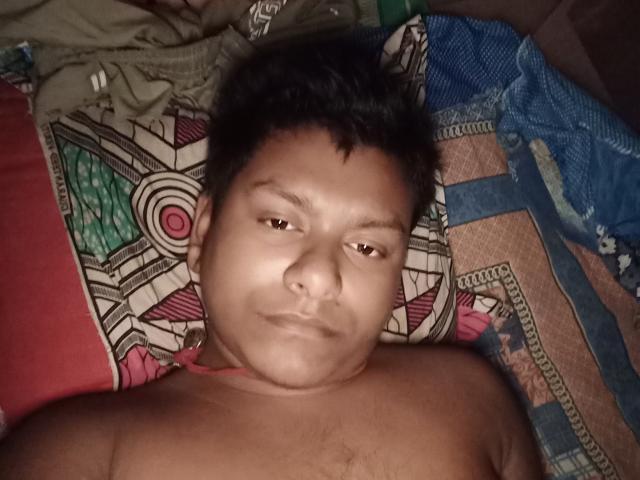 Shirtless SexyHotX69 with short black hair and brown eyes, lying on patterned bed, looking slightly to the side with a neutral expression, chest hair visible.