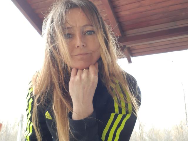MissFreeBird with messy blonde hair, blue eyes, and a slight smirk, wearing a black Adidas track jacket with yellow stripes, touching her chin.