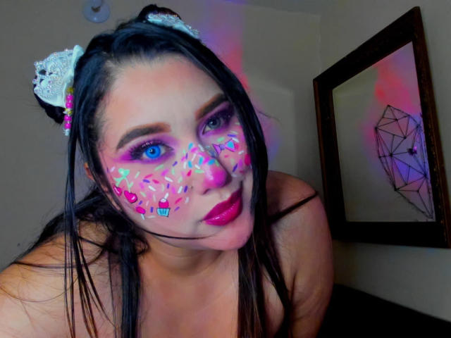 MadeleineDeveraux has glitter on her face, pink lipstick, and white cat ears, with colorful makeup dots. She has blue eyes, black hair, and is leaning forward, showing her tits.