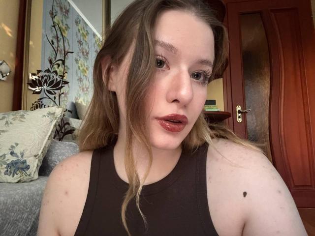 Tomioka69 has fair skin, brown eyes, and blonde hair. She's wearing a black tank top, showing her small, round tits with a black mole on her left shoulder.