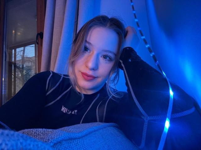 Tomioka69, con capelli biondi, occhi azzurri e rossetto rosso, indossa una camicia a righe nere, è sdraiato su un divano illuminato da una luce blu, con una mano tra i capelli e un leggero sorriso..