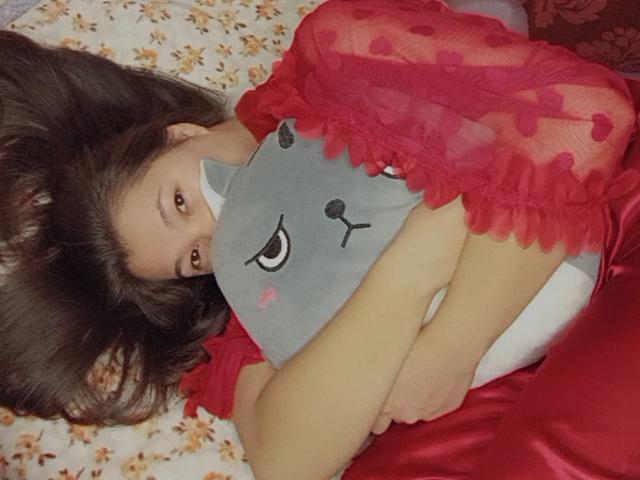 PeachyPalette lying on a bed, hugging a gray cat pillow, wearing a red lace dress with her tits partially covered, brown hair spread out, looking at the camera.