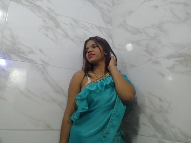 KairaDayamai with brown hair, red lipstick, wearing a blue ruffled dress, hand on neck, standing against white marble wall, looking slightly upward. Boobs partially covered.