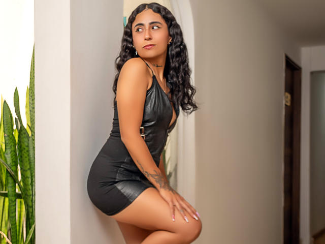 AriVelandia with curly black hair, wearing a tight black leather dress, sitting with her ass up and looking to the side, showing a tattoo on her right arm.