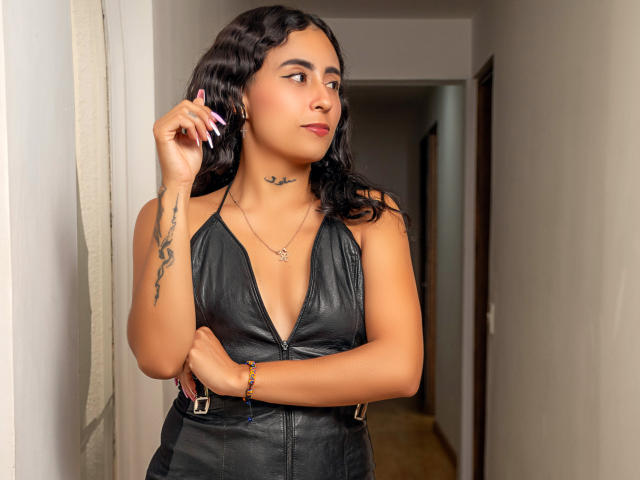 AriVelandia with curly black hair, tan skin, tattoos on arm and neck, wearing a black leather halter dress, standing in a hallway, looking to the side, touching her hair.