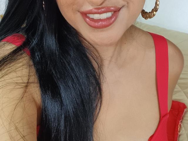 Apasion's shiny black hair, red lipstick, and red dress with a low neckline showing her tits. She's wearing gold hoop earrings, smiling slightly.