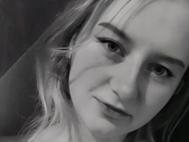 LolaMinnie's face, gray-scale, showing her small tits, slight smile, and long eyelashes, with messy blonde hair, looking directly at the camera.