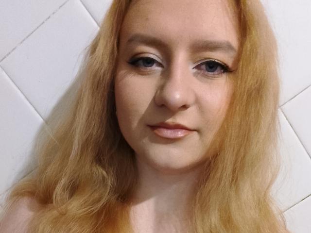LolaMinnie has blonde hair, blue eyes, and a cute face with a slight smile. She's looking directly at the camera with subtle makeup and a soft, natural look.