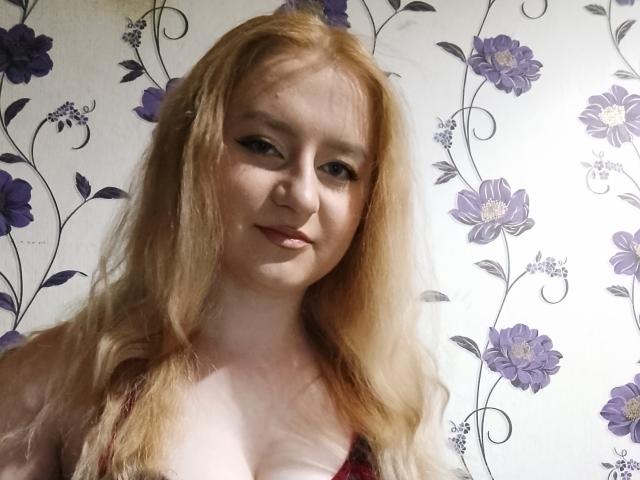 LolaMinnie with pale skin and long, blonde hair, wearing a red and black plaid top, showing small tits, smiling softly, looking at the camera.