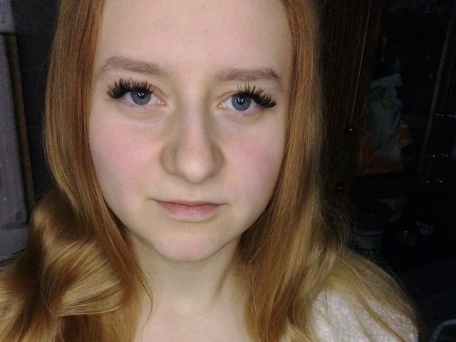 LolaMinnie has fair skin, red hair, blue eyes with long eyelashes, and a slight smile. Her small nose and subtle pink lips are visible. She's wearing a white top.