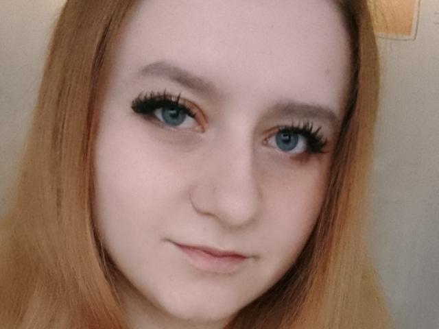 LolaMinnie has pale skin, blue eyes, and long red hair with thick black eyelashes. Her small, cute nose and subtle smile are visible. Her ass is not shown.