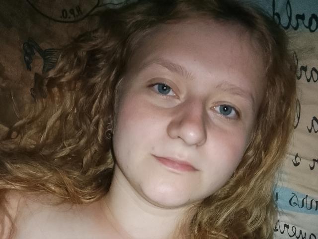 LolaMinnie has curly red hair, blue eyes, and a pale complexion. She's lying down, looking directly at the camera with a slight, relaxed smile. No clothes are visible.