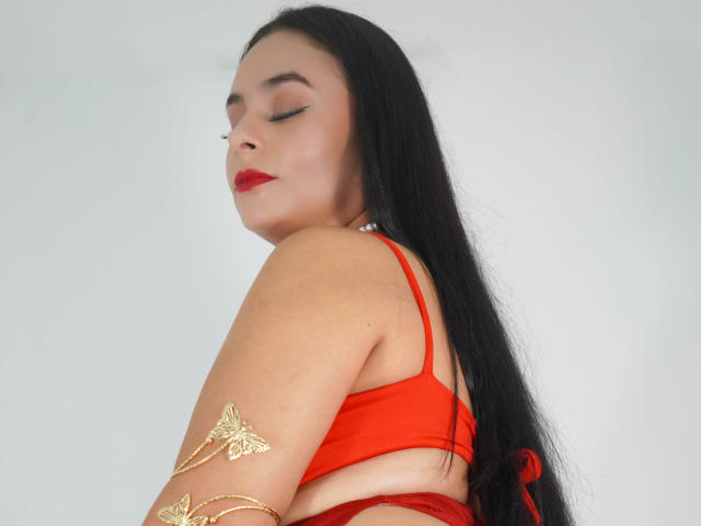 LunaSoler, con lunghi capelli neri, rossetto rosso e reggiseno rosso, occhi chiusi, con tatuaggi dorati sulle braccia, che si guarda oltre la spalla, con slip rossi visibili, su sfondo bianco..