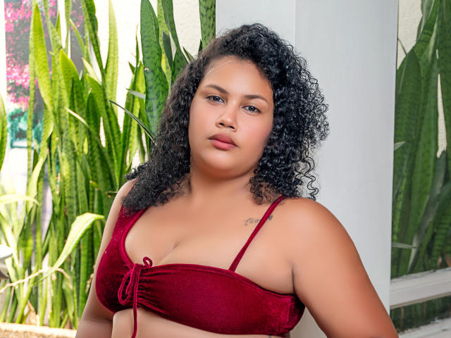 MiaGus, con i capelli neri ricci, la pelle marrone e piccoli seni, indossa un top di bikini rosso in velluto, è in piedi di fronte a piante verdi e guarda leggermente di lato..
