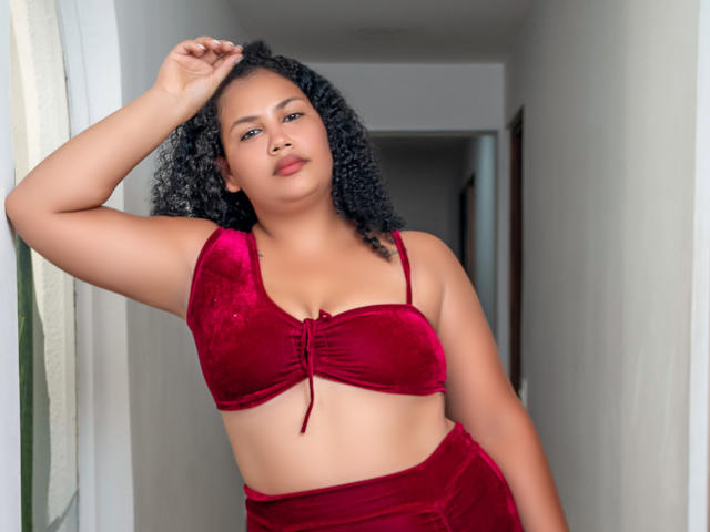 MiaGus, avec ses cheveux noirs bouclés, portant un soutien-gorge en velours rouge et un pantalon assorti à taille haute, se tient dans un couloir, une main dans ses cheveux, avec assurance et sensualité, peau brun moyen, petits seins..