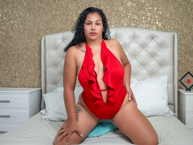 MiaGus, com cabelo preto encaracolado, pele marrom média, vestindo um maiô vermelho com babados e alças, sentada na cama, com as mãos nas coxas, mostrando sua vulva, nádegas e seios pequenos, com tatuagens..