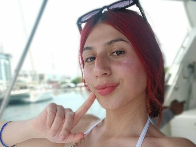 SophieBeaumont with red hair, wearing sunglasses on head, white halter top, pointing at slightly flushed face with finger, pink lips, small nose ring, blue bracelet, boat in background.