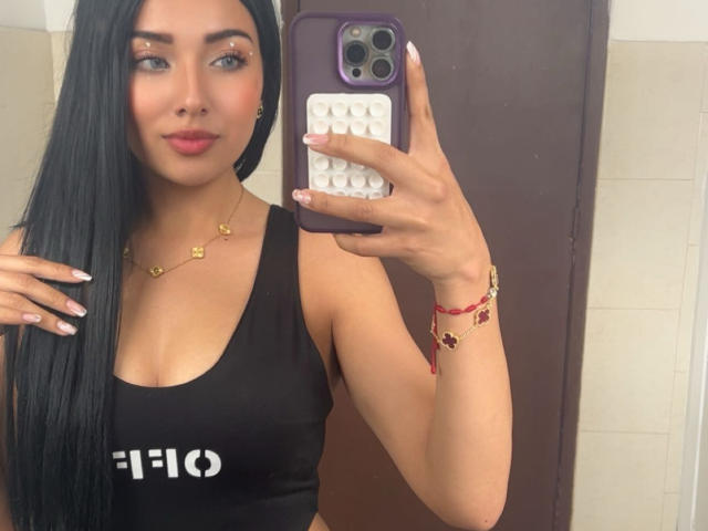 LillyTaylor with long black hair, wearing a black tank top, holding a purple phone with white textured case, gold necklace, red bracelet, pink lipstick, and looking at mirror.