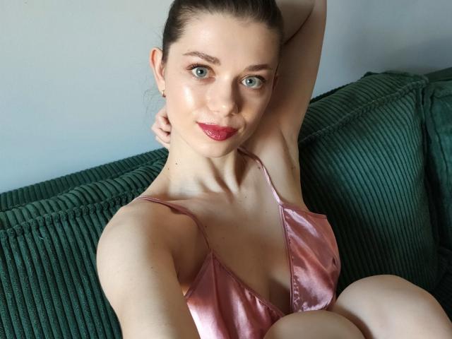 EvaMcCoy with pale skin, green eyes, and red lipstick, wearing a pink satin top, arm raised, sitting on a green ribbed couch, looking at the camera.