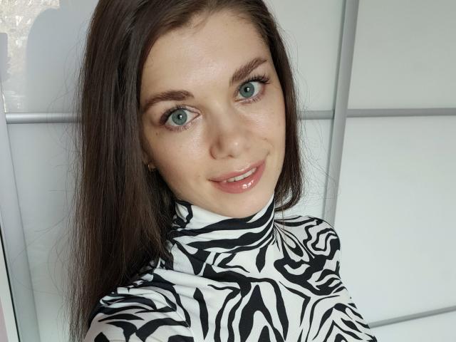 EvaMcCoy with green eyes, glossy pink lips, and brown hair, wearing a zebra-patterned turtleneck, smiling at the camera. Her tits are not visible.