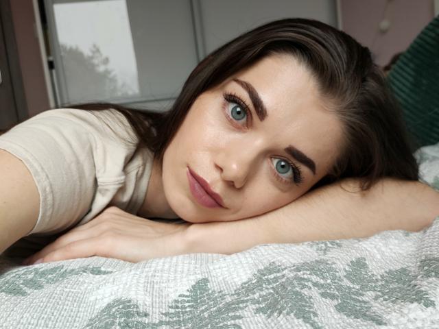 EvaMcCoy with green eyes, lying on a bed, head resting on arm, wearing a beige shirt, brown hair, pink lipstick, and subtle makeup, looking at the camera.