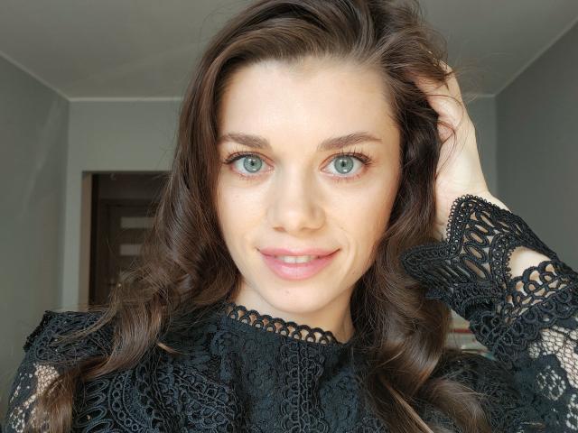 EvaMcCoy with green eyes, brown wavy hair, black lace top, smiling, hand in hair, looking at camera, soft natural light, subtle makeup, pink lips.