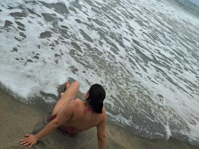 PaulaAnd with dark hair in a pink bikini, sitting on wet sand with her back to the camera, looking at foamy ocean waves. Her ass is partially visible.