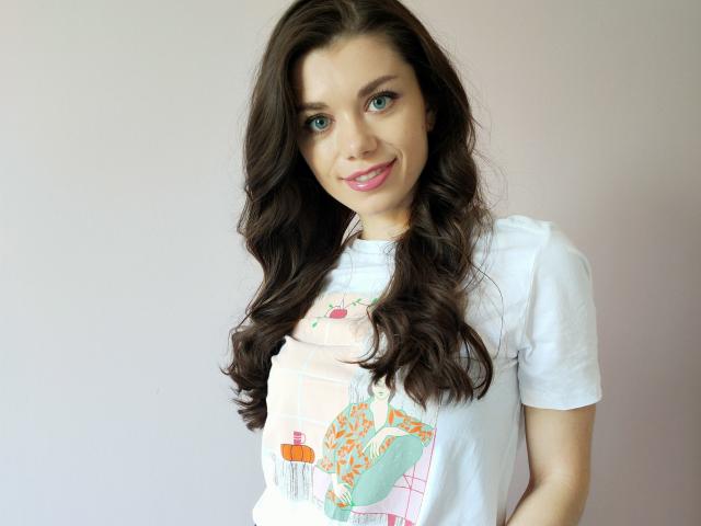 EvaMcCoy with long, wavy brown hair, green eyes, and pink lipstick, wearing a white t-shirt with a colorful print, smiling slightly. Boobs visible under shirt.