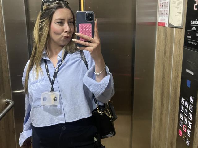 JennyLawry, con cabello rubio, camisa azul, pantalones negros y un distintivo de identificación, está tomando una selfie frente a un espejo dentro de un ascensor. Está haciendo pucheros, lleva gafas de sol en la cabeza y sostiene un teléfono rosa..