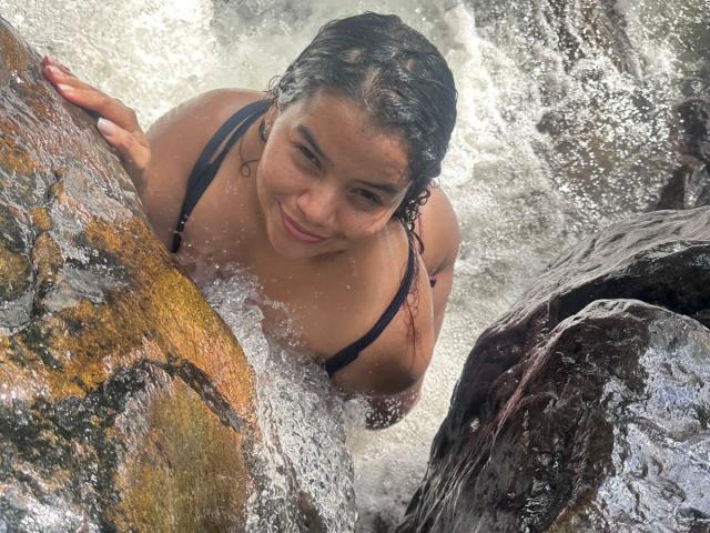 LolyMarce, wet with dark hair, smiling, leaning on rocks in a waterfall, wearing a black bikini top, her ass and tits visible, water splashing around.