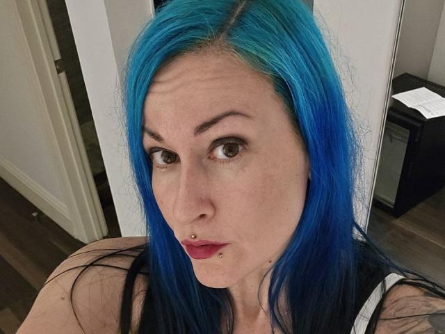 GennyRock, com cabelo azul vibrante, piercing no nariz e piercing no lábio inferior, olhando para a câmera com as sobrancelhas levantadas, vestindo uma blusa preta e branca..
