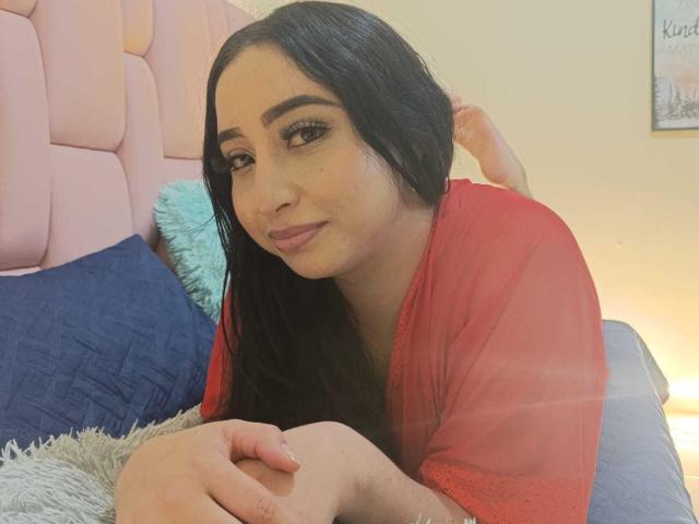 ValentinaVillar with long black hair, red sheer top, and subtle makeup, lying on a bed with a pink headboard, blue pillow, and grey blanket, looking at the camera.