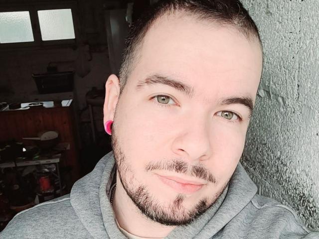 Transgenre69 hat einen rasierten Kopf, grüne Augen, pinke Kopfhörer, eine graue Kapuzenjacke und einen hellen Bart mit Schnurrbart und blickt mit einem leichten Lächeln in die Kamera..