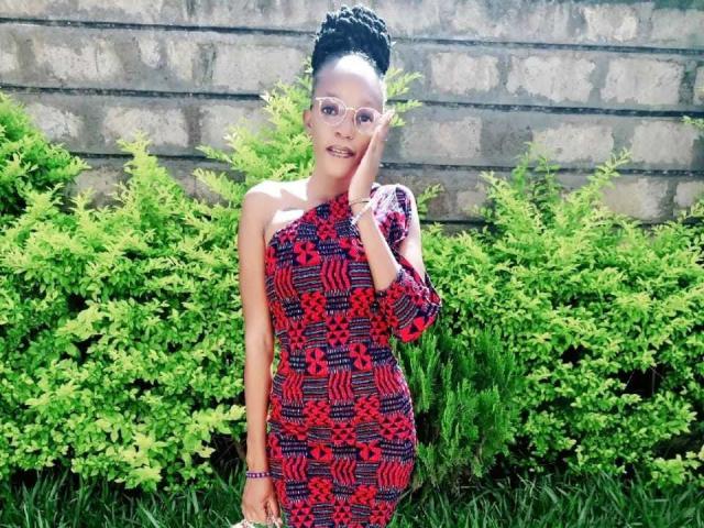 PrettyLoveHot with short black hair in a bun, wearing a red and blue patterned dress, glasses, and a bracelet, standing in front of green bushes, touching her cheek.