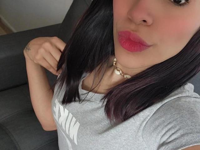 Madison Roberts, con rossetto viola scuro, maglietta grigia, capelli neri, un piccolo piercing al naso e una collana di perle, tiene i capelli dietro le orecchie, seduta su un divano grigio..