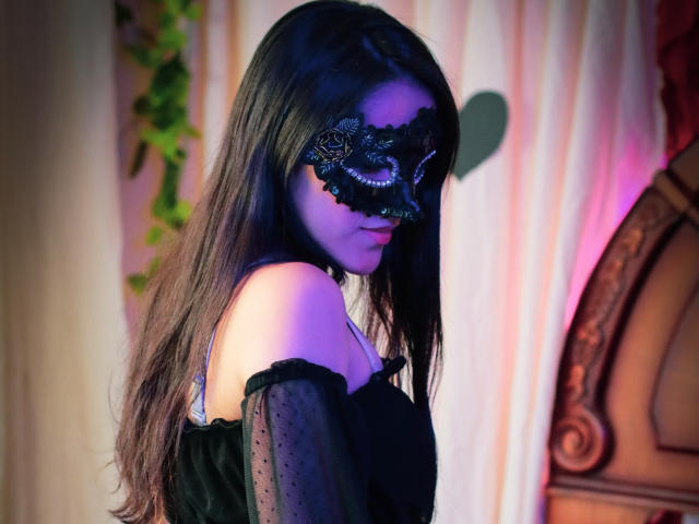 Loreh mit langem, dunklem Haar, einer schwarzen Maske und einem schwarzen Kleid mit abgesetzten Schultern, das ihre nackten Schultern und ein violettes Licht zeigt. Ihr Hintern ist teilweise sichtbar. Sie schaut nach unten und wirkt schüchtern..