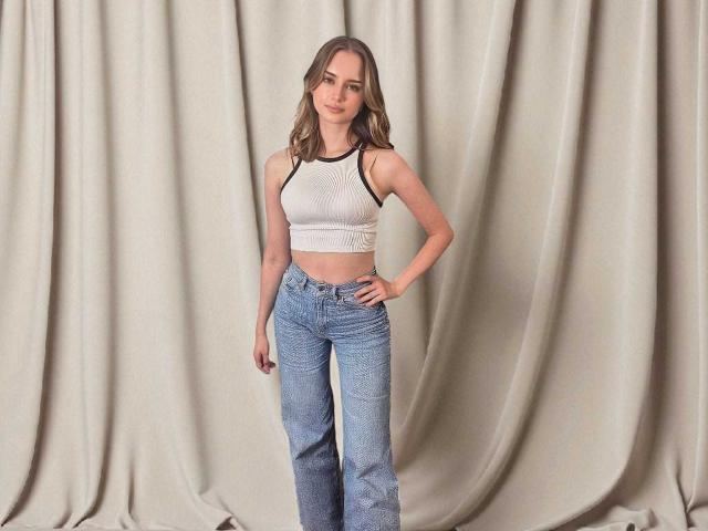 GraceKissable with wavy brown hair, white sports bra showing her small tits, and high-waisted blue jeans, standing against beige draped curtains, hand on hip, smiling.