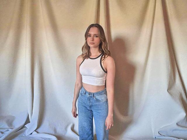 GraceKissable stands with wavy brown hair, wearing a white sleeveless top and high-waisted blue jeans, showing her small tits and flat ass, against a beige draped background.