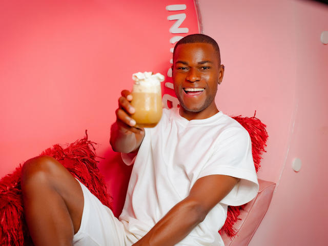 JakeHottest, a dark-skinned dude with a shaved head, is smiling while holding a frothy drink, wearing a white tee and shorts, sitting on red pillows.