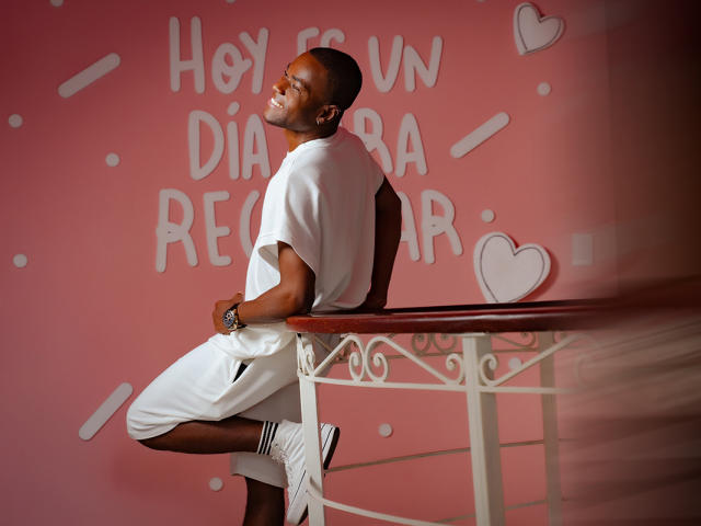 JakeHottest, with dark skin and a short beard, leans on a white table in a white outfit, smiling, showing his ass, and wearing white sneakers.