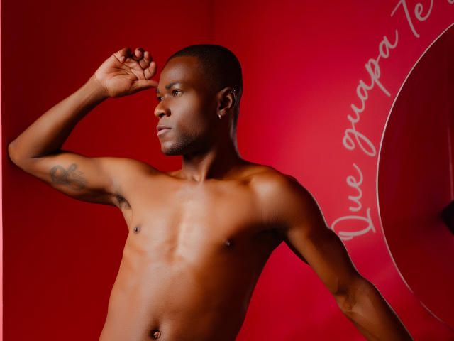 Shirtless JakeHottest with dark skin, short hair, small nipple piercings, tattoo on right arm, and a confident pose against a red background. He's looking to the side with a slight smile.