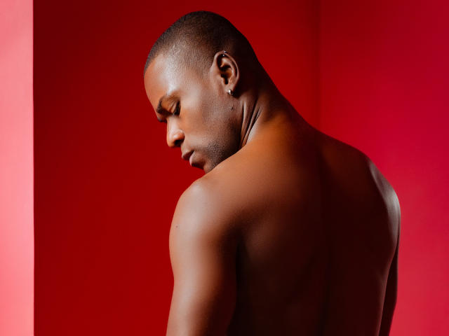 JakeHottest, shirtless, with dark skin, short buzz cut, looking down, showing muscular back, red background, smooth skin, ear with small hoop earring, serious expression, upper body shot.