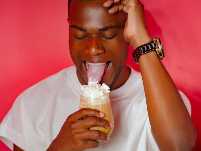 JakeHottest with dark skin, shaved head, white t-shirt, and wristwatch, licking a creamy, whipped-topping dessert with his tongue out, holding it with one hand against a pink background.