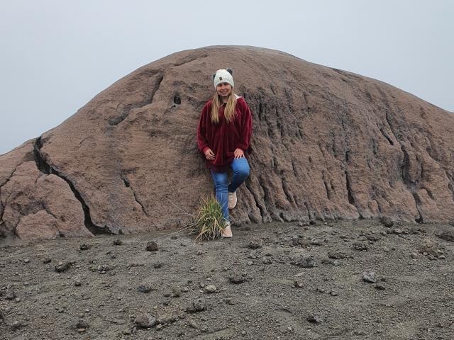 LucianaSeashell está em frente a uma grande rocha, vestindo um gorro branco, um casaco de veludo vermelho, jeans azuis e sandálias, com grama na mão direita, sorrindo..