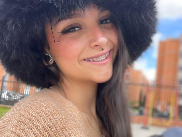 MarietteJones with a black furry hat, nose piercings, and a smile showing metal braces. She's wearing a textured beige sweater and black circular earrings.