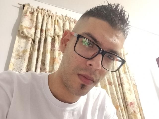 GolFather has short, spiked black hair, wearing black glasses and a white tee, with a goatee, looking slightly down at the camera. His face is slightly flushed.