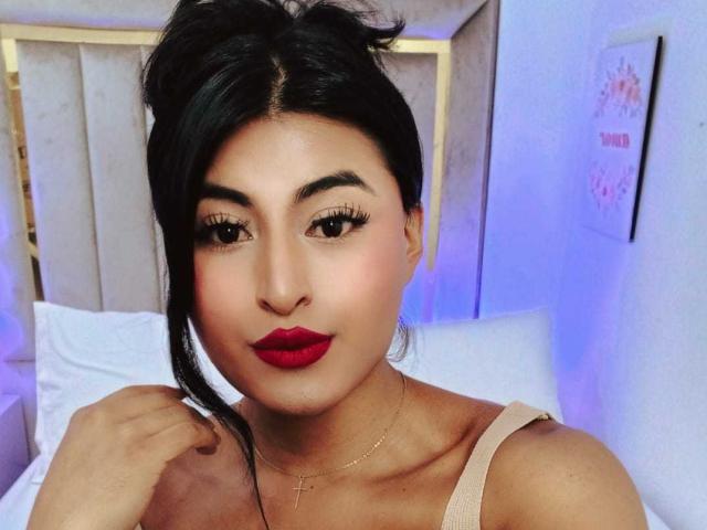 MichelleDenise with dark hair in a messy updo, red lipstick, gold cross necklace, and beige top, showing off her tits and smooth, tan skin in a bedroom.