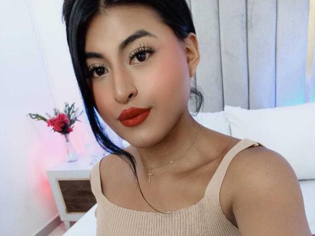 MichelleDenise with tan skin, black hair, red lipstick, cross necklace, and beige ribbed tank top showing her tits. Her ass is not visible. Bedroom background with flowers.